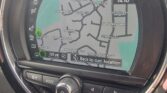 A MINI's GPS navigation screen shows a map with surrounding streets, including High Street, Oxenden Road, and London Road. There is a selection button for returning to the current location. The screen is surrounded by various control buttons and knobs. Time displayed is 14:10.