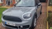 A grey MINI Cooper with the license plate FV67 YSY is parked on a brick driveway in front of a red brick house with white-framed windows and a black door. The driveway extends to a detached garage in the background. There is a small green lawn and hedge to the left side of the car.