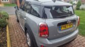 A gray MINI Cooper Countryman is parked on a brick driveway in front of a residential area. The car boasts a yellow license plate with the registration number "FV67 YSY." Brick houses and neatly trimmed lawns with small shrubs create a charming backdrop.