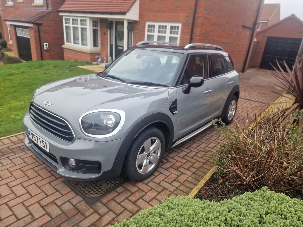 A silver MINI Countryman SUV is parked in a brick driveway in front of a red-brick house. The car has two doors on each side, silver alloy wheels, and a black roof with roof rails. The background includes a green lawn, bushes, and another house with a red tile roof.