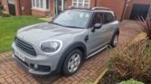 A silver MINI Countryman SUV is parked in a brick driveway in front of a red-brick house. The car has two doors on each side, silver alloy wheels, and a black roof with roof rails. The background includes a green lawn, bushes, and another house with a red tile roof.