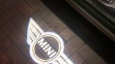 A close-up photo showcases the illuminated MINI logo projection on a brick pavement. The logo, featuring the word "MINI" inside a circular design with wings extending outward, is also reflected on the side of a car in the upper part of the image.