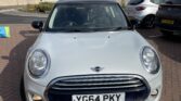 A silver MINI Cooper is parked on a brick pavement. Its trunk is open, revealing the interior. The license plate reads "YG64 PKY." There are other cars and a high concrete retaining wall in the background with some trees and a streetlight visible at the top of the wall.