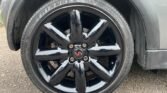 A close-up image of a shiny, black car wheel with a slick six-spoke design. The tire has visible tread patterns and branding that reads "Triangle." The car body is metallic gray, reflecting the classic style of a MINI. The ground appears rough and textured. A person is partially visible in the background on the left side.