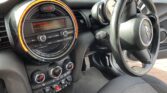The interior of the MINI car features the steering wheel on the right side and a circular infotainment system in the center. The screen displays a radio station with buttons below. Air conditioning controls and various switches are located beneath. The background showcases a brick wall.