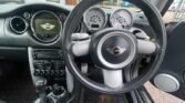 Wide-angle view of the interior of a Mini Cooper. The dashboard features a large circular speedometer and smaller gauges, with the iconic MINI logo on the steering wheel. The center console has numerous controls, and the gear shift is visible at the bottom left.