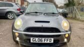 A gray MINI Cooper with headlights illuminated is parked on a gravel driveway, with houses in the background. The license plate reads "GL06 FFD." The photo is taken from the front, showcasing the car's grille, hood scoop, and fog lights. Trees and a chain-link fence are visible on the right.