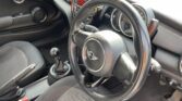Interior view of a MINI car focusing on the driver's seat and dashboard. Features include a black leather-wrapped steering wheel with the Mini logo, a manual gear stick, round air vents, and a compact, clean dashboard with a speedometer and control buttons. Upholstery is black with fabric seats.
