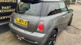 A gray MINI Cooper S with black alloy wheels is parked on a gravel surface. The vehicle, bearing a UK license plate "GL06 FFD," features a dual exhaust system and a rear spoiler. The car is next to a building with signs offering warranties, part exchange, and services.