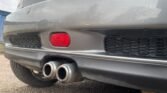 Close-up of the rear end of a gray MINI. The image prominently features the car's dual exhaust pipes, a red reflector, and a black mesh grille on the rear bumper. The surroundings include a portion of the asphalt ground and the lower part of a blue structure in the background.