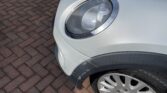 Close-up of a light-colored MINI's front left side, showing a minor dent and scratched area near the headlight. The car is parked on a brick-paved surface. The image highlights the headlight, the damaged spot, and the front wheel with a tire.