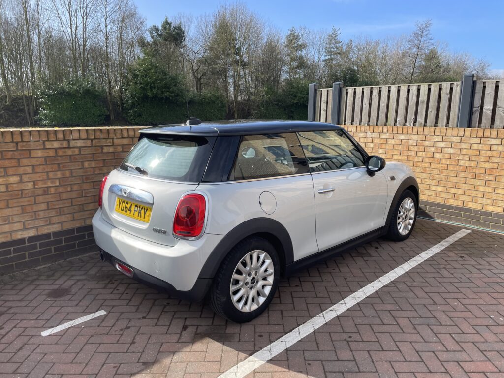 A silver MINI Cooper with a black roof is parked in a designated space facing a brick wall. The license plate reads "VO64 FKY." The car is positioned near a wooden fence and surrounded by trees in the background under a clear sky.