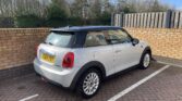 A silver MINI Cooper with a black roof is parked in a designated space facing a brick wall. The license plate reads "VO64 FKY." The car is positioned near a wooden fence and surrounded by trees in the background under a clear sky.