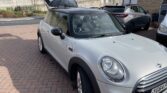 A silver MINI Cooper with a black roof is parked on a brick driveway. The car's rear hatch is open, revealing the spacious cargo area. The license plate reads "YG64 ZVJ." In the background, there are other parked cars, a building, and a large concrete wall with visible bushes and trees.