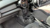 The interior of this compact MINI car showcases the driver's side. The dashboard features a central circular display, air vents, and climate control dials. The center console includes a gearshift, cup holders, and handbrake. The steering wheel has mounted controls, while the seats are upholstered in dark fabric.