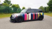 A black MINI Cooper with a vibrant, multicolored stripe design on the body is parked on a paved road. The car has a low stance, black rims, and tinted windows. Trees and green foliage line the background, partially hidden by wire fencing, under an overcast sky.