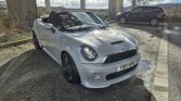 A white MINI Cooper S convertible with its top down is parked under a bridge on a gravel path. The car has black alloy wheels and a visible license plate reading "YB61 NPG." Another black SUV is parked in the background, and a cloudy sky looms in the distance.