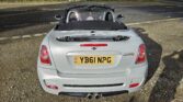 A rear view of a silver MINI Cooper S convertible car with a UK license plate reading "YB61 NPG." The car's top is down, revealing a two-seater interior with headrests. The MINI is parked on a gravel surface near a divided road, with grass and shadows visible in the background.