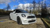A white MINI Cooper is parked outdoors in a sunlit area with a bright blue sky and scattered clouds overhead. It is positioned on a gravel surface next to a road. The car has black wheels, a sunroof, and a license plate reading "YB61 WPG." Leafless trees and a wire fence are in the background.