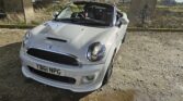 A silver MINI convertible is parked on a gravel road under a concrete bridge. The car's top is down, revealing black leather seats. The license plate reads "YB61 NPG." Sunlight casts shadows on the ground, and a grassy field with a fence is visible in the background.