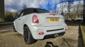 A silver convertible MINI Cooper with a black soft top is parked on a gravel surface. The vehicle's UK license plate reads "YB61 NPC". The car has black wheels and dual exhaust pipes. In the background, there are trees and a building under a partly cloudy sky.