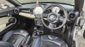 Interior of a modern compact MINI car featuring a black and beige color scheme. A steering wheel with a branded logo, an analog speedometer, and a tachometer dominate the dashboard. The center console has control knobs and buttons. Two black leather seats with beige accents are visible.