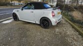 A silver MINI convertible car with a black soft top is parked on a gravel surface next to a curb. It has black alloy wheels and a visible rear license plate with the registration "YC62 TSY." The car is positioned facing away from the camera, with trees and a road in the background.
