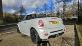 A white MINI convertible car with a UK license plate reading "V861 WPG" is parked on a gravel surface. The vehicle boasts black alloy wheels and an open top. In the background, a partly cloudy sky and bare trees hint at a chilly yet sunny day.