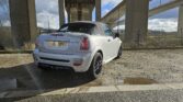 A white MINI convertible with black rims is parked under a bridge on a gravel surface. The vehicle is facing away from the camera, showcasing its rear and license plate. The sky is partly cloudy, and the environment features concrete pillars and a chain-link fence.