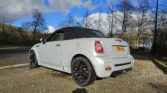A white MINI convertible with a black soft top roof is parked on a gravel surface against a backdrop of leafless trees and a blue sky with scattered clouds. The car has a UK license plate, black alloy wheels, and dual exhaust. The sun casts shadows, indicating it's a bright, clear day.