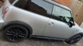 A silver MINI Cooper with black alloy wheels is parked on a driveway next to a brick wall and a wooden gate. The car has tinted windows, a black roof, and visible reflections on the glass. The rear hatch and tail light are visible, as well as part of the car's interior through the windows.