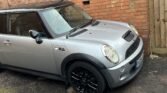 A silver MINI Cooper with a black roof is parked on a slightly sloped driveway next to a brick house. The car features black alloy wheels and a hood scoop. In the background, there's a wooden fence to the right and an exterior wall of the house with a drainpipe.