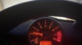 A MINI's tachometer glowing with red lighting displays an engine speed of around 9 RPM x 1000. Below the tachometer, a small digital screen shows the temperature as 24.3 degrees Celsius. The steering wheel is partially visible, and a garage door is seen through the windshield.