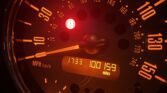 Close-up of a MINI speedometer illuminated in red and orange light. The dial shows speeds up to 150 mph or 240 km/h. The digital display reads "17:33" and "100159 miles." A circular red warning light is lit near the 20 mph mark, representing a car dashboard at night.