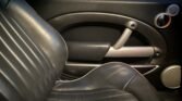 A close-up view of a black leather car seat and door interior, showcasing the signature MINI design. The door features an oval handle encased in silver trim, accompanied by a round speaker. A set of car keys rests on the seat while part of a brick wall is visible through the window.