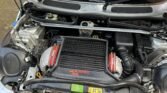 The image shows the engine bay of a MINI with the hood raised, revealing an engine equipped with an Artec-branded component. The engine area appears clean and slightly wet, possibly due to rain. Various engine parts, wires, and fluid reservoirs are visible around the central engine block.