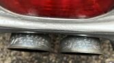 Close-up view of a MINI's dual exhaust pipes and rear light assembly with raindrops on the surface. The metal exhaust tips have "WORKS" etched onto them. In the background, wet pavement with small pebbles is visible.