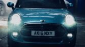 A teal MINI Cooper with license plate AK16 NXY is parked head-on under a light source. The car's headlights are on, illuminating the wet ground and surrounding area. A white car is partially visible in the background. The scene appears to be an indoor garage or car wash.