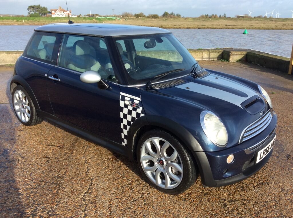 A dark blue MINI Cooper with racing stripes and a checkered flag pattern on its side is parked on a wet gravel surface next to a large body of water. Wind turbines and some trees can be seen in the background. The license plate reads "L852". The car has alloy wheels and a sunroof.