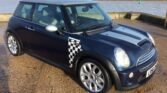 A dark blue MINI Cooper with racing stripes and a checkered flag pattern on its side is parked on a wet gravel surface next to a large body of water. Wind turbines and some trees can be seen in the background. The license plate reads "L852". The car has alloy wheels and a sunroof.