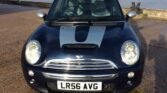 A dark-colored MINI Cooper with a silver racing stripe on the hood is parked on a gravel surface near a body of water. The car's front view includes round headlights, a chrome grille, and the license plate number "LR56 AVG." The background features a concrete barrier and some greenery.