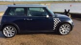 A parked MINI Cooper with a black body and white checkered design on the side is by a waterfront. The car features silver rims and a distinctive roofline. In the background, there is a body of water, grassy areas, and wind turbines on the horizon. The sky is partly cloudy.