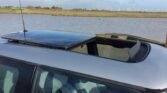 A silver MINI with an open sunroof is parked near a water body under a partly cloudy sky. In the background, there are wind turbines along the horizon and a small green buoy floating in the water. The interior of the car is partially visible through the sunroof.