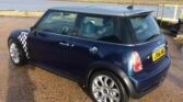 A blue MINI Cooper S is parked on a paved area near a body of water. The car features a British racing flag pattern on its front fender and sports alloy wheels. The weather appears cloudy with the sun breaking through. A house and tree line can be seen in the background across the water.