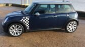 A dark blue MINI Cooper with a black and white checkered decal on the side is parked on a gravel surface near a body of water. Wind turbines stand tall in the background on grassy land. The car has silver alloy wheels and a shiny, clean exterior.