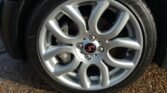 Close-up view of a silver alloy wheel rim with a distinctive swirl design, fitted with a black tire. The center cap features a red "S" in a black circle, signature to MINI. The wheel is part of the car parked on a pebbled surface.