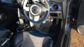 The interior of the car features a blue and silver steering wheel with the iconic MINI logo in the center. The dashboard includes several gauges and control buttons. The car boasts two-tone leather seats in black and silver, a manual transmission, and an open driver's door revealing the audio system.