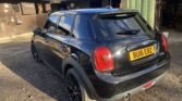 A black MINI Cooper with license plate BU16 EBZ is parked on a gravel surface beside a green building and stables. The car's glossy exterior reflects sunlight, with its rear and side windows darkly tinted. The background includes various objects and a "Sonney" sign on the stable.