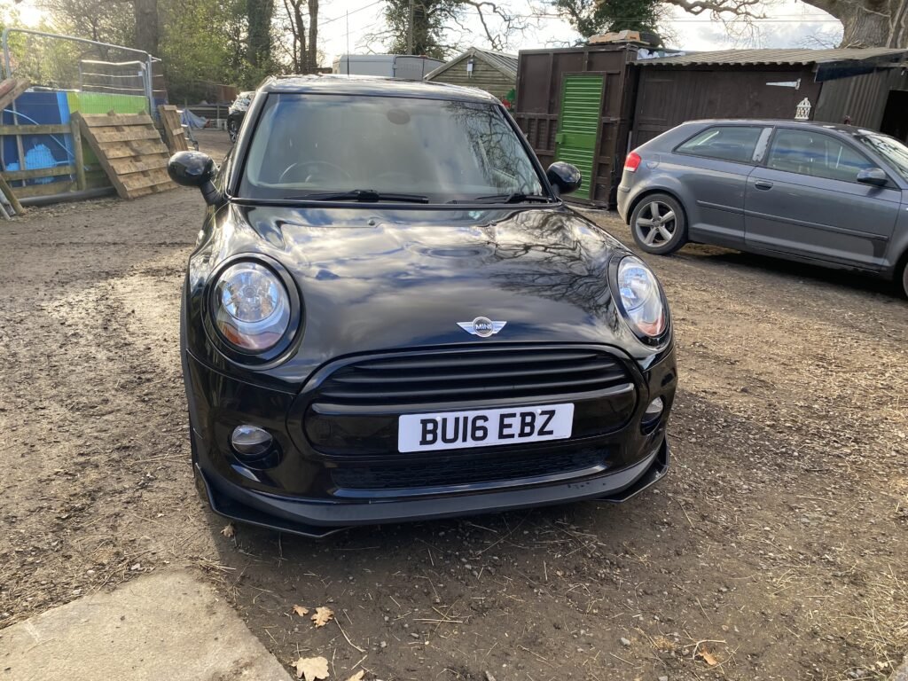 A black MINI Cooper with the registration plate "BU16 EBZ" is parked on a dirt surface. Another car, silver in color, is parked to the right. There are various sheds, trees, and fencing in the background, creating a rustic and semi-urban setting.