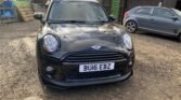 A black MINI Cooper with the registration plate "BU16 EBZ" is parked on a dirt surface. Another car, silver in color, is parked to the right. There are various sheds, trees, and fencing in the background, creating a rustic and semi-urban setting.